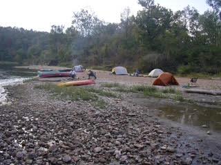Tents and canoes on a wide gravel bar beside Bryant Creek at dusk, with wooded hills behind