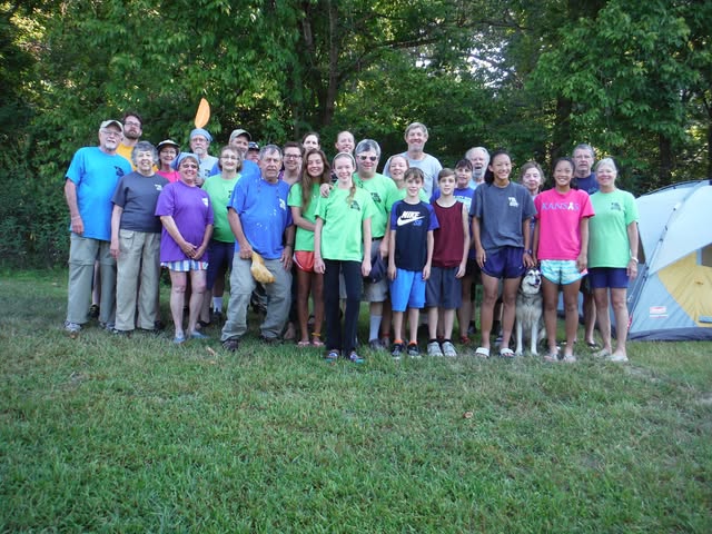 About thirty OWWC members of all ages, many in matching green club shirts, posing together at a wooded campsite next to a tent