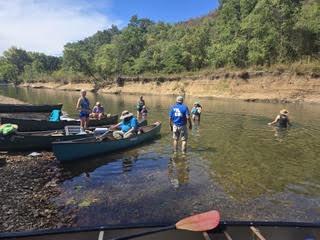 Club members wading beside loaded green canoes in shallow, clear water below a wooded bank